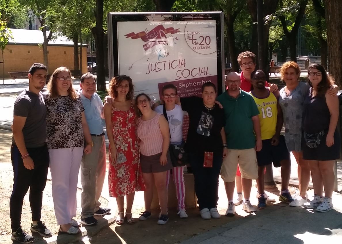Foto de un grupo de personas pertenecientes al Área de Vida Adulta de la Asociación AMP posando delante de una cartel del evento II Feria de la Justicia Social